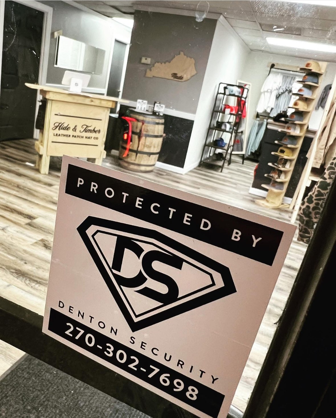 Glass storefront door of a local hat shop with a Protected by Denton Security sign showing the phone number 270-302-7698